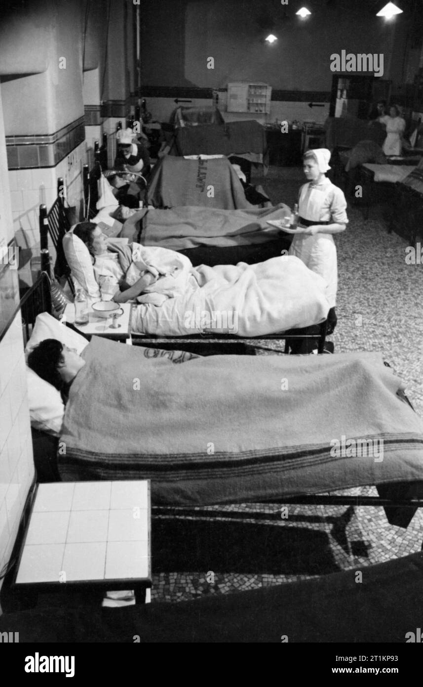 Krankenschwestern bei der Arbeit auf einer Station im Krankenhaus von Guy, London, 1941. Einen weiten Blick über das Leben auf der Station am Guy's Hospital in den frühen Morgenstunden. Eine Krankenschwester trägt ein Fach der Arzneimittel für einen Patienten, während eine andere Krankenschwester im Hintergrund feeds einen Patienten (mit einem großen Rahmen über ihre Beine) mit einem langen spouted Cup. Stockfoto