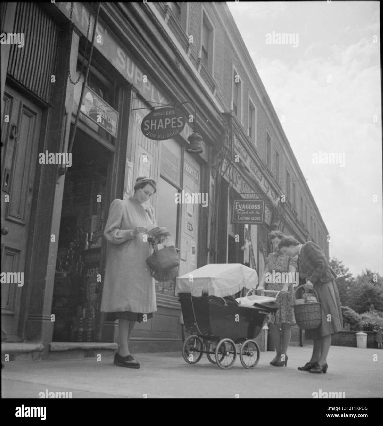 Alle - im Krieg Arbeiter - Alltag für Frau M Hasler, Barnes, Surrey, 1942 Frau Hasler, Warenkörbe in der Hand, überprüft Ihr bezugsschein als verlässt sie ein Geschäft. Zwei andere Frauen können in der Fotografie außerhalb dieses kleine Reihe von Geschäften gesehen werden: das eine ist eine junge Mutter mit einem Kinderwagen, der andere ist mit einem Korb und bewundern Sie das Baby. Stockfoto