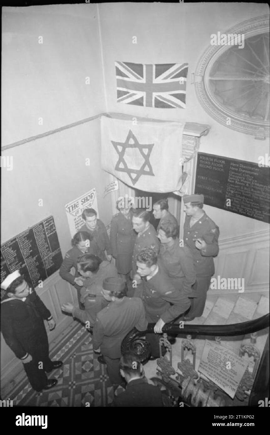 Die alliierten Streitkräfte Feiern das Jüdische Neue Jahr - religiöse Feiern im Balfour Service Club, London, UK, 1943 eine Gruppe von alliierten Soldaten und Soldatinnen in den Flur an der Balfour Service Club Menge, einige stehend auf der Treppe, die Rosch Haschanah feiern. Auf den Tafeln auf der linken Seite sind die Namen aller, die das Fest besucht geweißt. Gemäß der ursprünglichen Bildunterschrift, viele Menschen waren in der Lage, alte Freunde bei den Feierlichkeiten zu erfüllen. Eine Union Flag und eine Flagge mit dem Davidstern hängen an der Wand über dem Kopf. Stockfoto