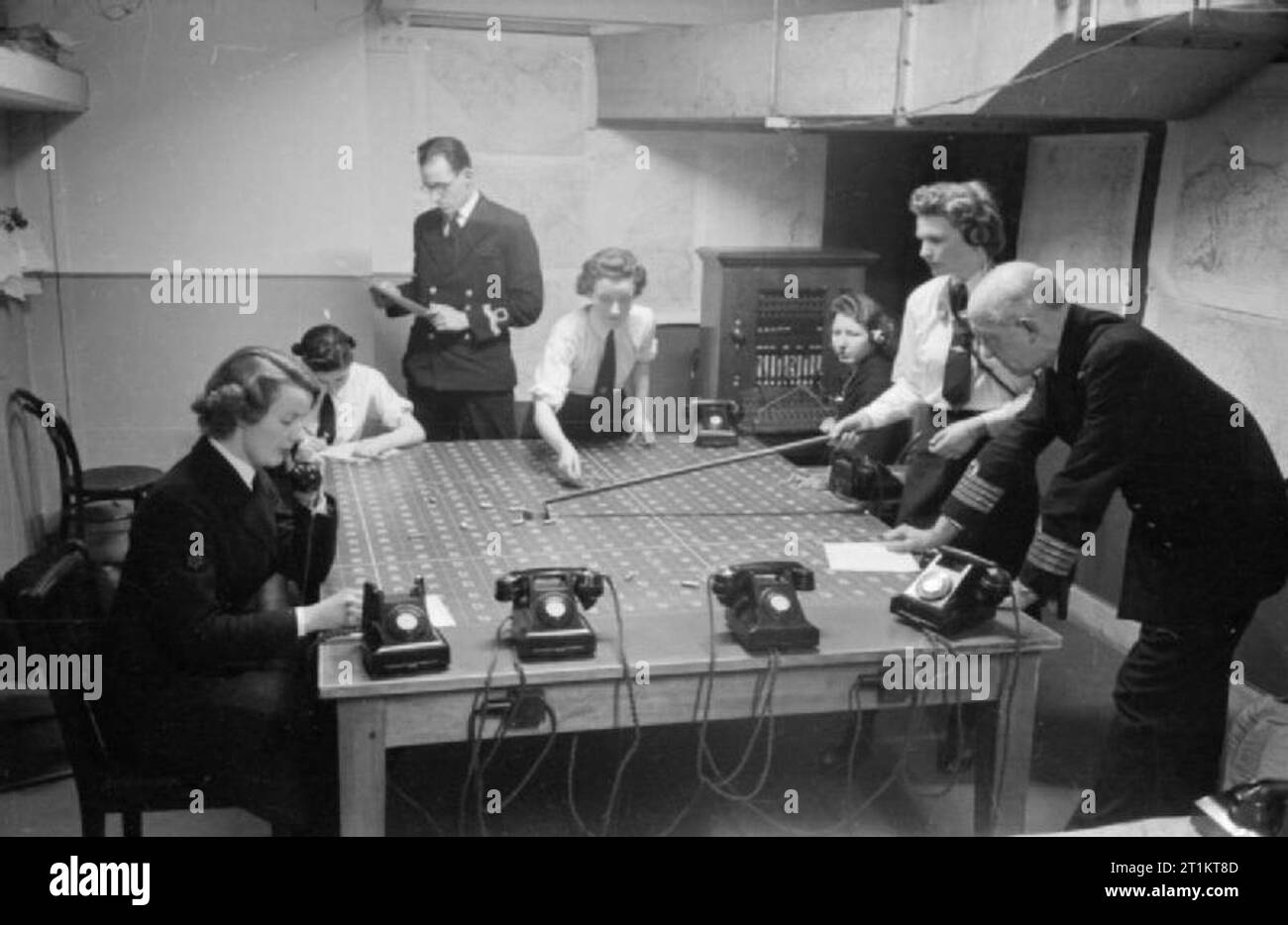 Das Unbekannte Mädchen hinter dem Meer Schlacht - die Arbeit von Women's Royal Naval Service, 1942 Mit einem Rechen, Wren Unbekannt bewegt ein "Schiff" über die nummerierten Grid Map auf dem Plotten Tabelle der Operationen Zimmer an HMS ADLER. Der Leiter der Personal steht zu Ihrer Linken, auf der rechten Seite des Fotos, und Uhren die Aktion entfalten. Mehr Zaunkönige bei der Arbeit rund um die Tabelle zu sehen sein, darunter eines über das Telefon und ein Mitglied der Royal Navy kann auch im Hintergrund zu sehen. Stockfoto