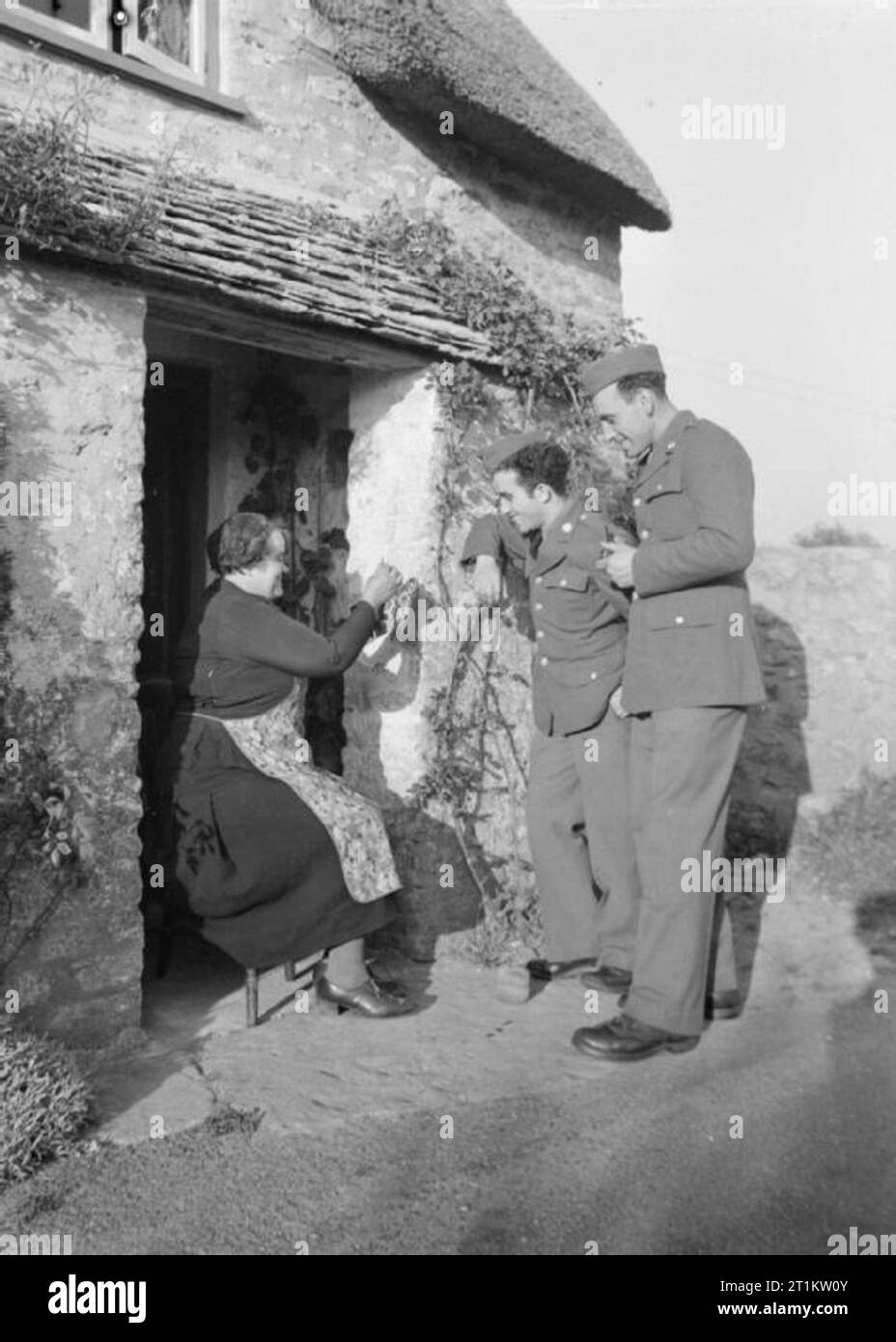 US-Truppen in einem englischen Dorf - den Alltag mit den Amerikanern im Burton Bradstock, Dorset, England, UK, 1944 amerikanische Soldaten zu Frau Guyatt vor ihrem Häuschen auf dem Weg zu ihrem Camp bei Burton Bradstock, Dorset zu chatten. T/4 Johnny Mandaro (von 3740 77th Street Jackson Höhen, Long Island, New York) und T/5 Samuel Goodman (von 534 Ritner Street, Philadelphia, Pennsylvania) zusehen, wie Frau Guyatt Zöpfe Netze in der Sonne. Stockfoto