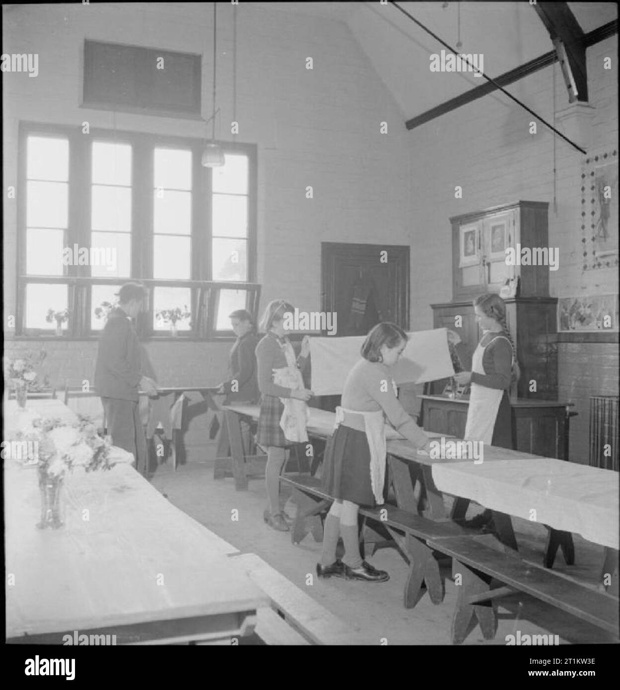 Gärten Feed Schulkinder - Nahrungsmittelproduktion auf Knighton-auf-Haus, Worcestershire, England, UK, 1943 Nach dem Mittagessen in der Dorfschule in Knighton - Haus der Frau, der Speisesaal ist gelöscht und zu seinem normalen Zustand als Klassenzimmer zurück. Jungen bewegen Sie die Bänke und Tische, während Mädchen, falten sich die Tischdecken. Stockfoto