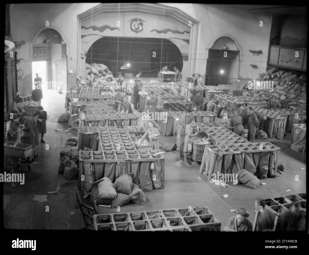 Army Post Office - Korrespondenz und E-Mail in Kriegszeiten, England, Großbritannien, 1944 eine belebte Szene in einer stillgelegten Dance Hall, irgendwo in den Midlands, die jetzt als die Armee Post verwendet wird. Die Sortierung wird derzeit von den Mitgliedern der Auxiliary Territorial Service. Mail wird zuerst in den verschiedenen Branchen der Dienstleistungen und dann sortiert, von dort in die einzelnen Einheiten sortiert ist. Große Netze mit Säcke verwendet werden die Briefe und Pakete in ihre verschiedenen Gruppen zu sortieren. Die Bühne und Arch im Hintergrund dienen als eine starke Erinnerung an die früheren Leben des Gebäudes. Stockfoto