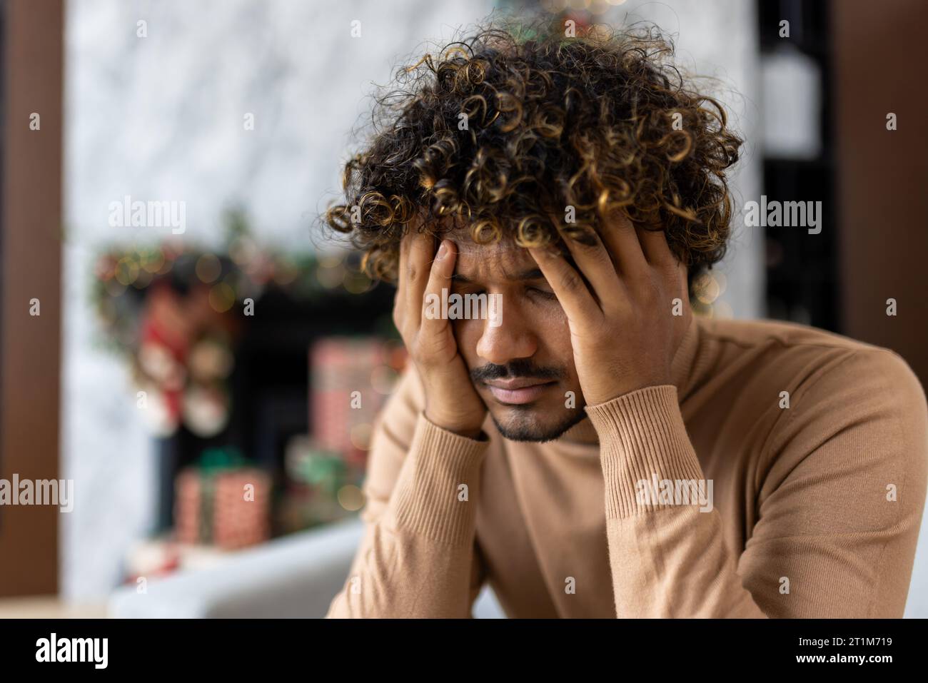 Starke Kater Kopfschmerzen und Migräne, Mann mit Kopf sitzend auf Sofa im Wohnzimmer zu Weihnachten, afroamerikanischer Mann mit lockigen Haaren in der Nähe von dekoriertem Weihnachtsbaum. Stockfoto