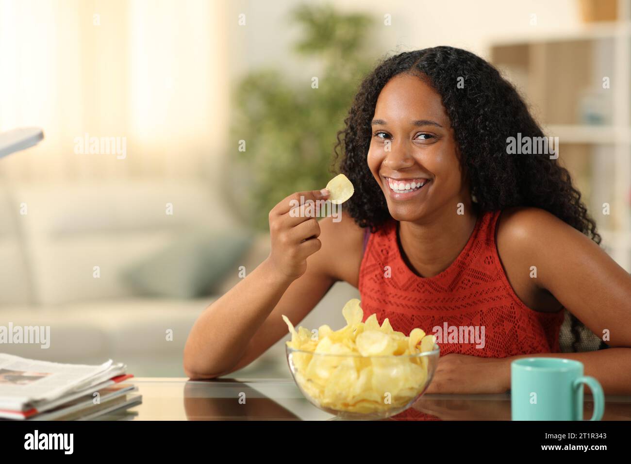 Glückliche schwarze Frau isst Kartoffelchips und posiert dich zu Hause an Stockfoto