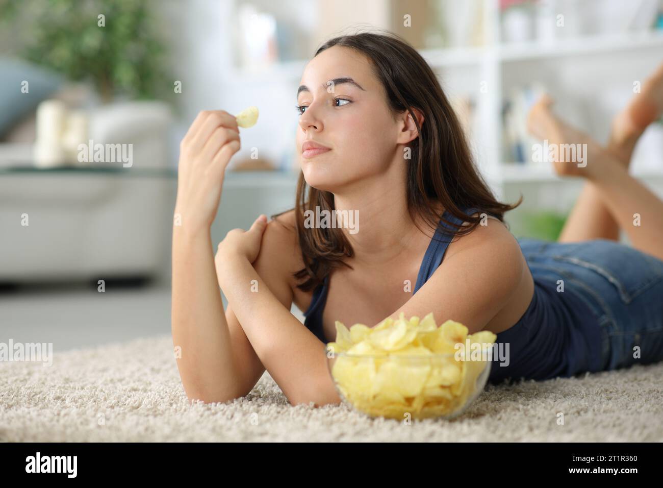 Nachdenkliche Frau, die wegschaut und Kartoffelchips isst, die zu Hause auf dem Boden liegen Stockfoto