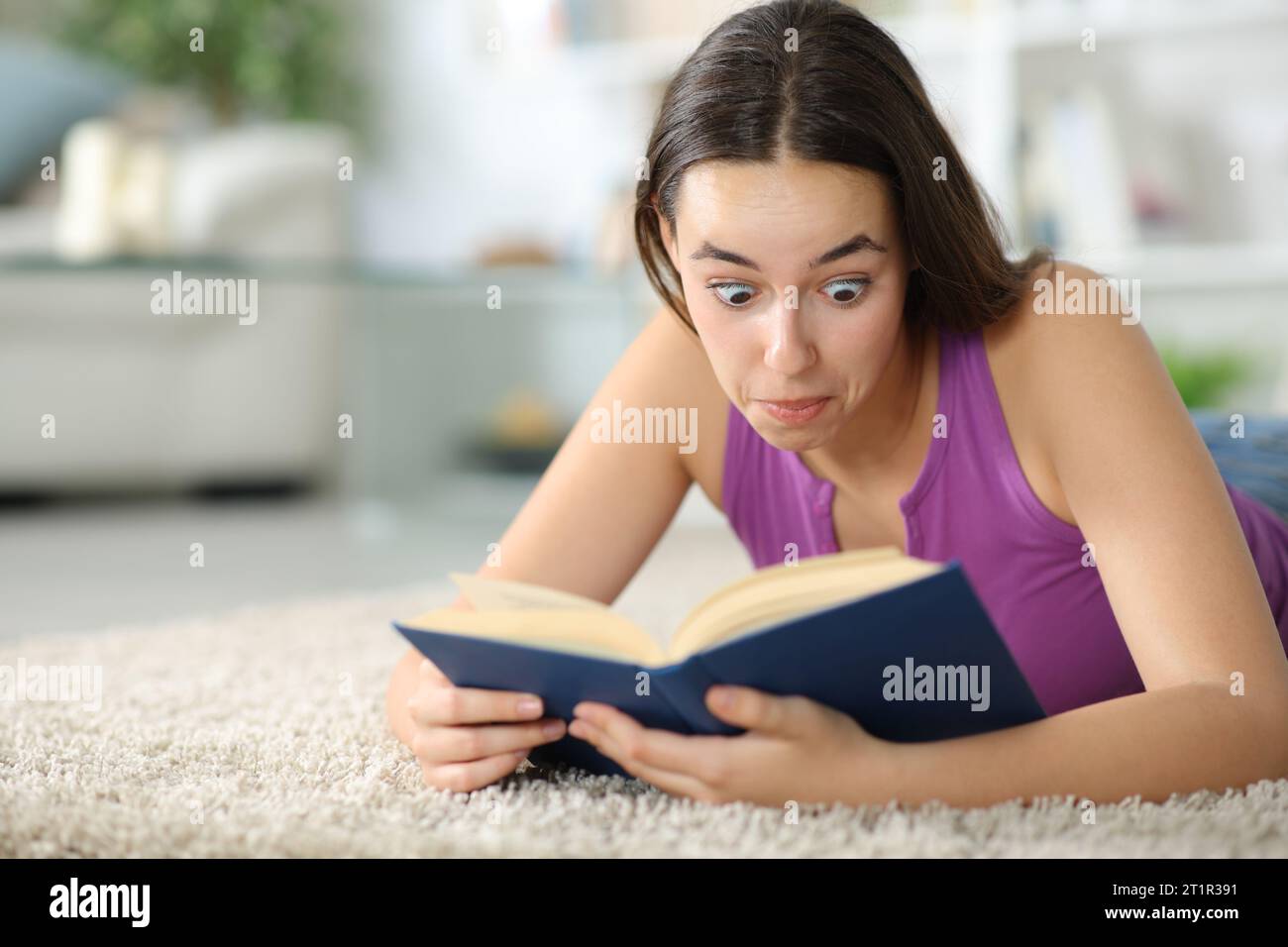 Überraschte Frau, die zu Hause ein Papierbuch liest, das auf einem Teppich liegt Stockfoto