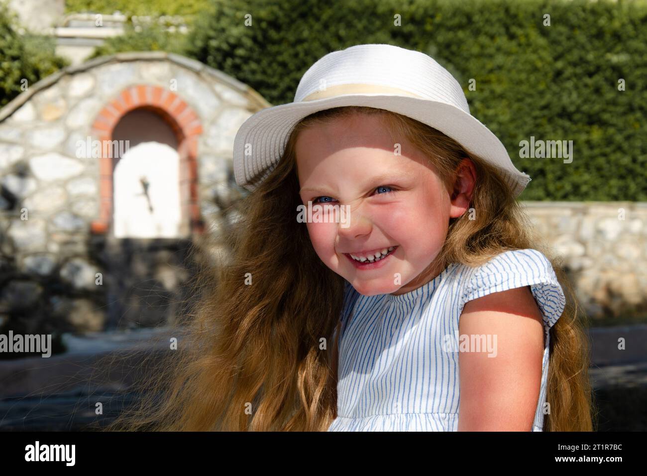 Ein Mädchen von etwa fünf Jahren in einer weißen Mütze mit lockeren blonden Haaren lacht überrascht. Porträt von der Taille nach oben. Weißes Mauerwerk und Grün in t Stockfoto