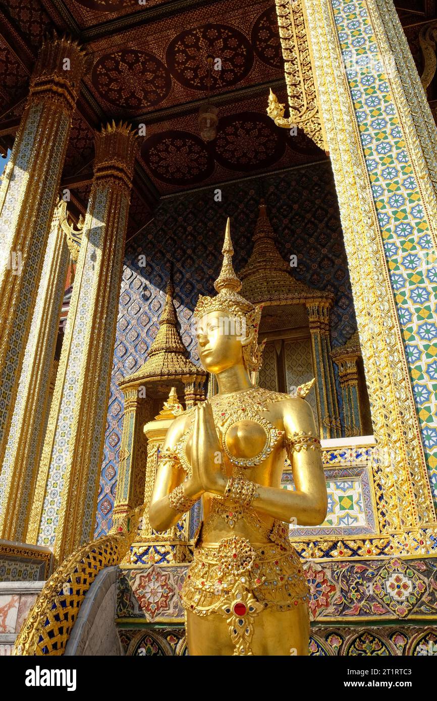 Ein Tempel mit komplexen blauen und goldenen Mosaikfliesen dient als Kulisse für eine goldene Statue einer Frau in traditioneller thailändischer Kleidung. Stockfoto
