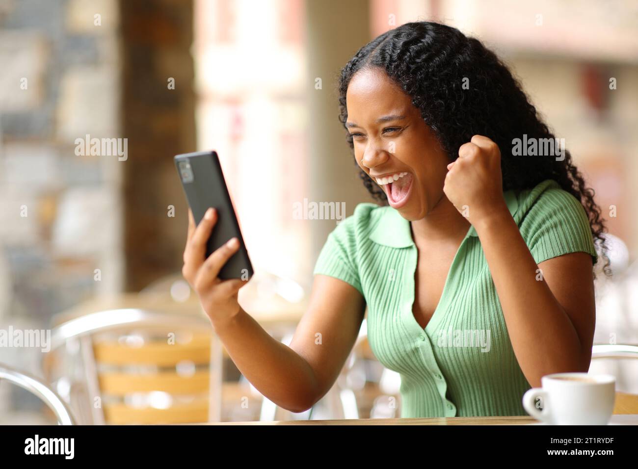 Aufgeregte schwarze Frau, die ihr Smartphone auf einer Bar-Terrasse überprüft Stockfoto