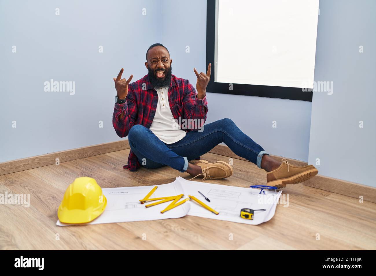 afroamerikaner sitzt auf dem Boden in einem neuen Zuhause und sieht sich Blaupausen an, die mit verrücktem Ausdruck schreien und Rock-Symbol mit erhobenen Händen machen. Musikstar Stockfoto