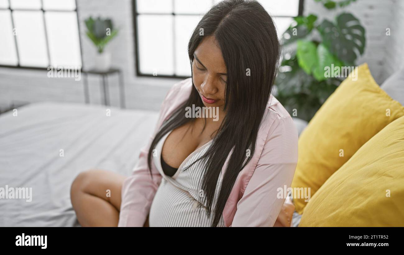 Eine junge schwangere Frau, die sich der Mutterschaft anschließt, sitzt entspannt auf dem Bett, blickt ernsthaft auf ihren Bauch in ihrem gemütlichen Schlafzimmer und denkt über den Morgen nach Stockfoto