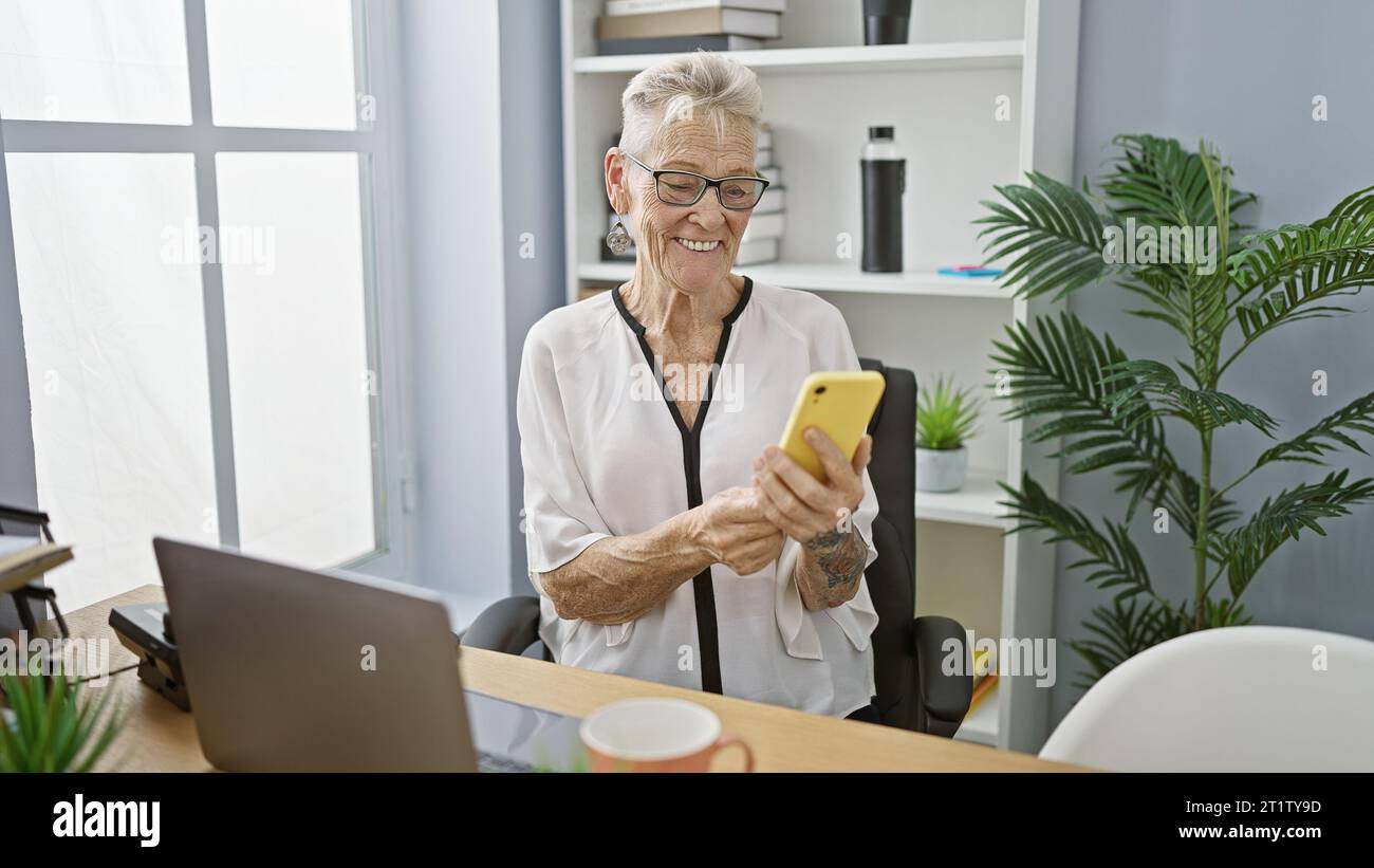 Lächelnde ältere grauhaarige Geschäftsfrau, die im Büro eine fröhliche Nachricht auf ihrem Smartphone schreibt Stockfoto