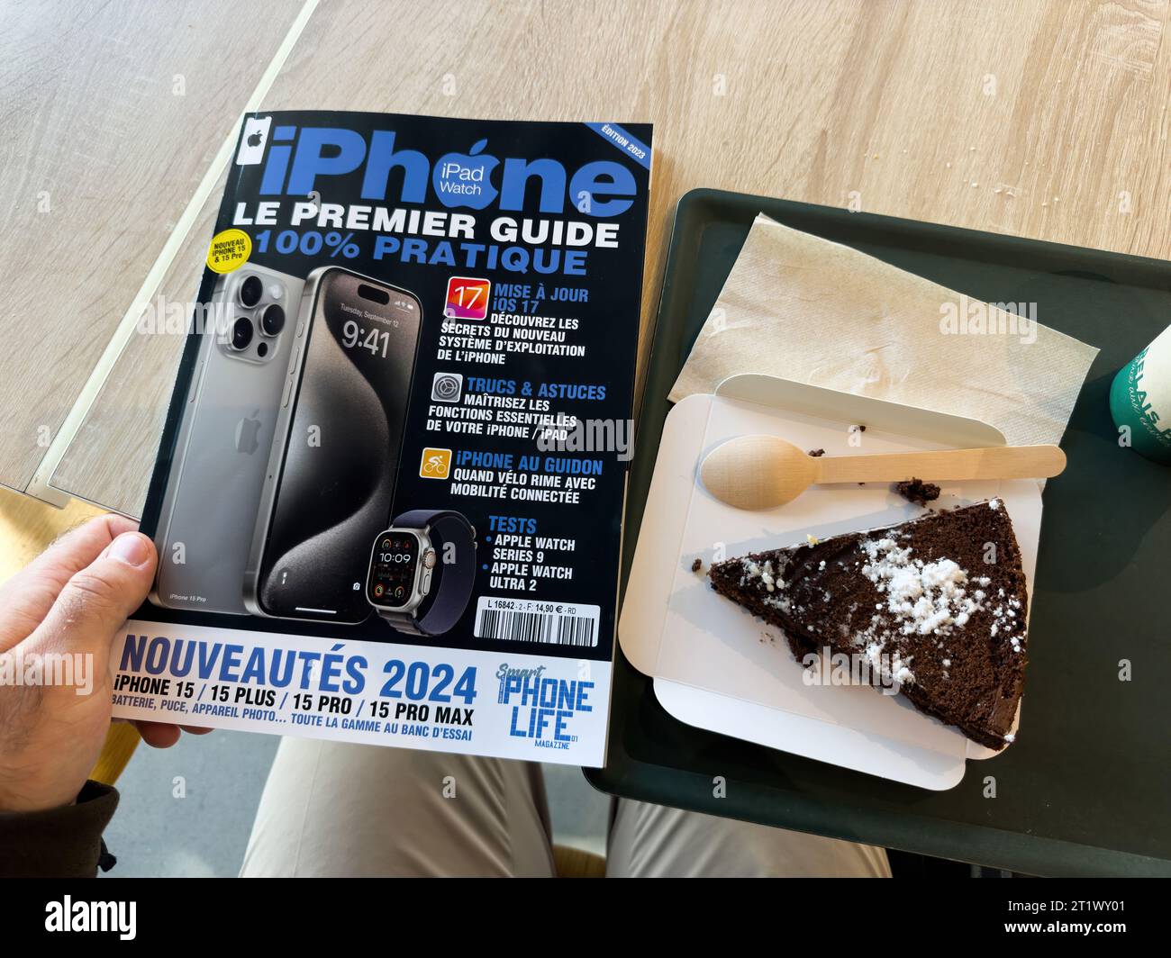 Paris Frankreich - 10. Oktober 2023: Männliche Hand hält iPhone das erste praktische Guide Magazin mit Ankündigung von 2024 und dem neuesten 15 Pro Modell neben einem Café-Schokoladenkuchen Stockfoto