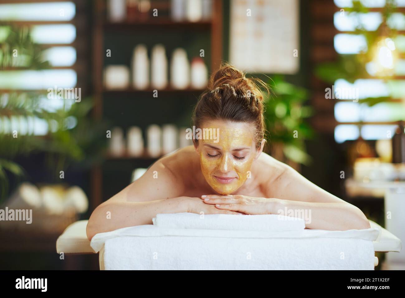 Gesundheitszeit. Entspannte, moderne 40-jährige Frau im Spa-Salon mit goldener Kosmetikmaske auf dem Gesicht, die auf einem Massagetisch liegt. Stockfoto