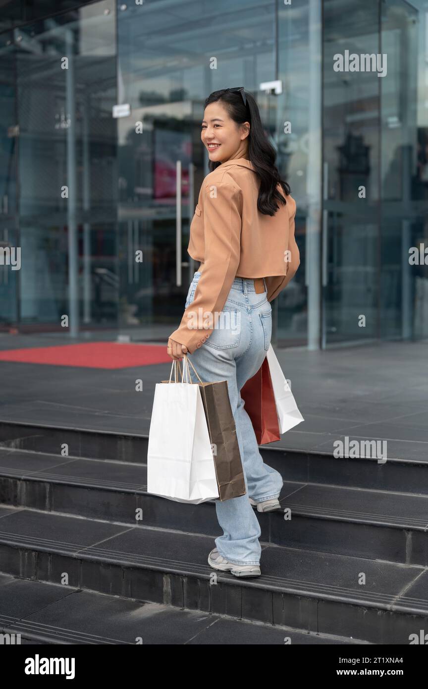 Eine fröhliche und schöne junge asiatische Frau in trendigen Kleidern geht in ein Einkaufszentrum, geht mit ihren Einkaufstaschen und genießt ihr Einkaufen Stockfoto