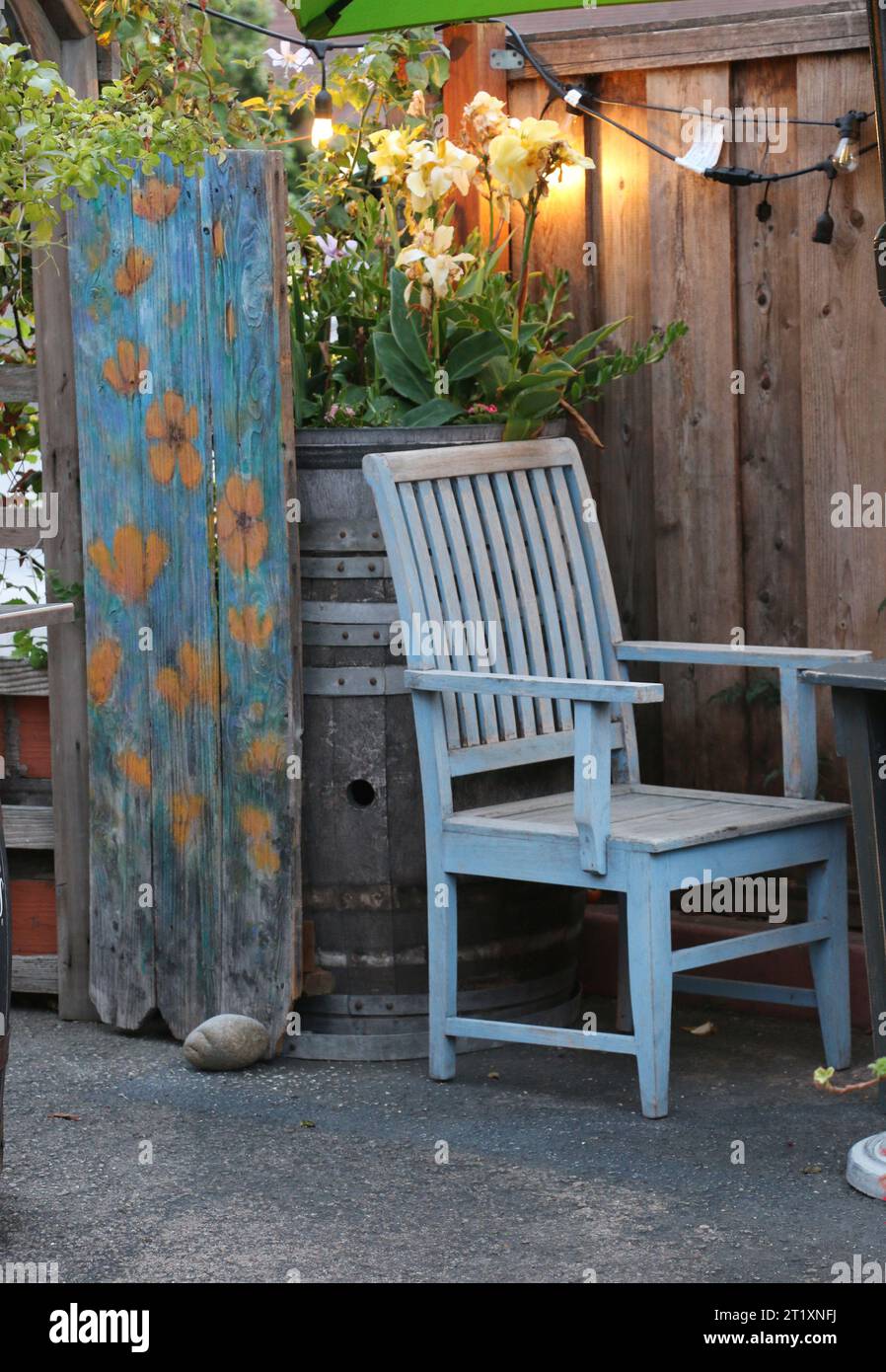 Ein verwittertes Tor, blau mit gelben Blumen bemalt, dient als einladender Eingang zu einem Gartenrestaurant-Innenhof. Ein verwitterter blauer Stuhl steht in der Nähe. Stockfoto