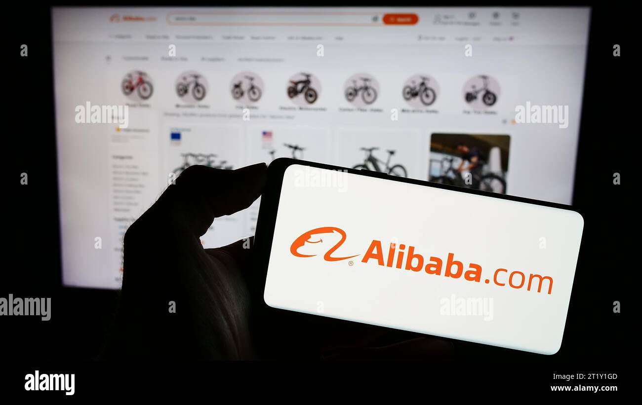 Person, die ein Mobiltelefon mit dem Logo der chinesischen B2B-E-Commerce-Plattform Alibaba.com vor der Business-Website hält. Schwerpunkt auf dem Display des Telefons. Stockfoto