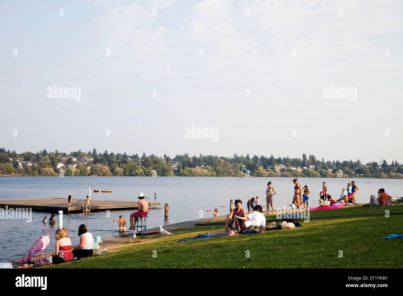 Die Menschen sitzen in der Sonne am Ufer eines Sees, während die Menschen sich sonnen und im Hintergrund schwimmen. Stockfoto