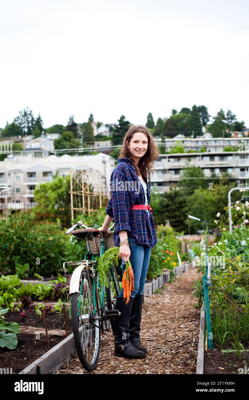 Eine junge Frau lächelt und blickt in die Kamera, während sie neben ihrem Fahrrad steht und ein Bündel Karotten hält. Sie ist eine von vielen, die sich in der städtischen Landwirtschaft engagieren und sich täglich darum kümmern Stockfoto