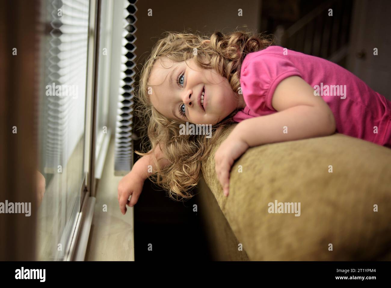 Kleinkind mit blonden Locken und blauen Augen, die lächelnd aus dem Fenster schauen Stockfoto