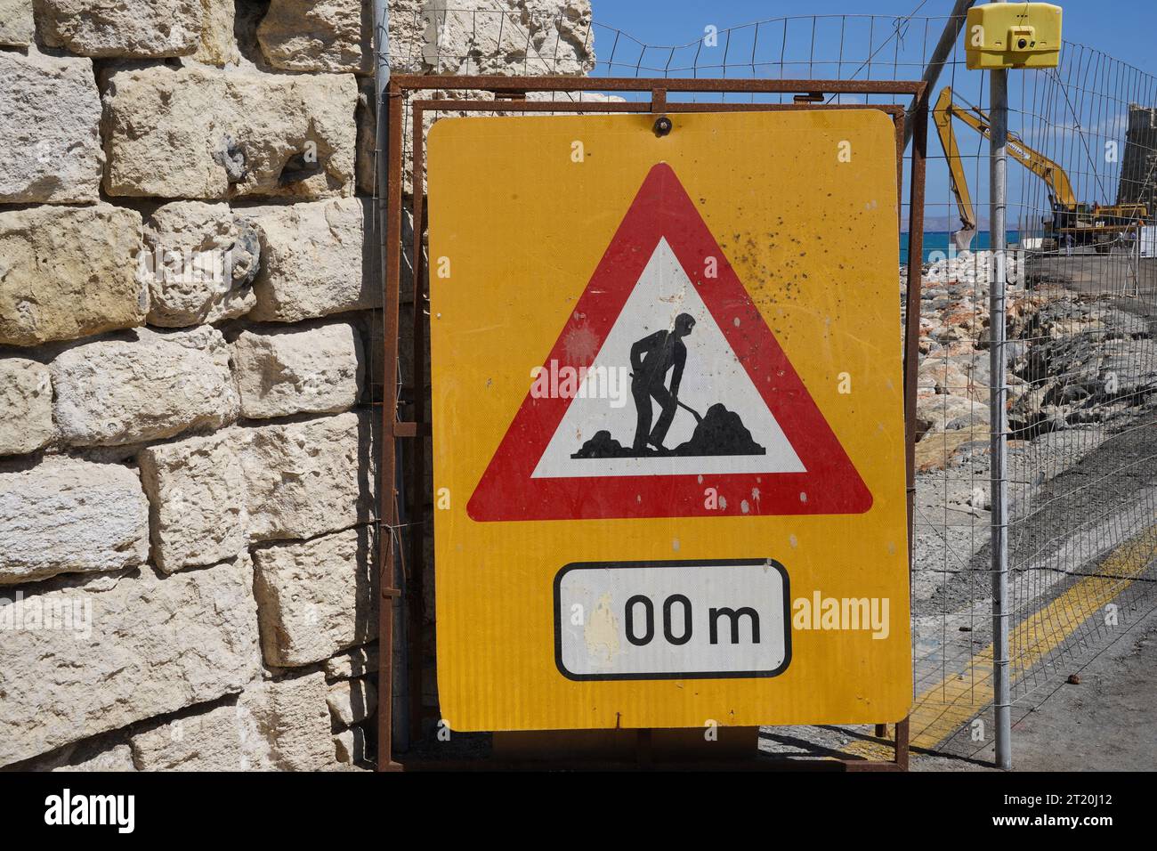 Dreieck-Verkehrsschild Men at Work auf einem gelben Metallschild. Dahinter befindet sich blaues Meer und Bagger. Stockfoto