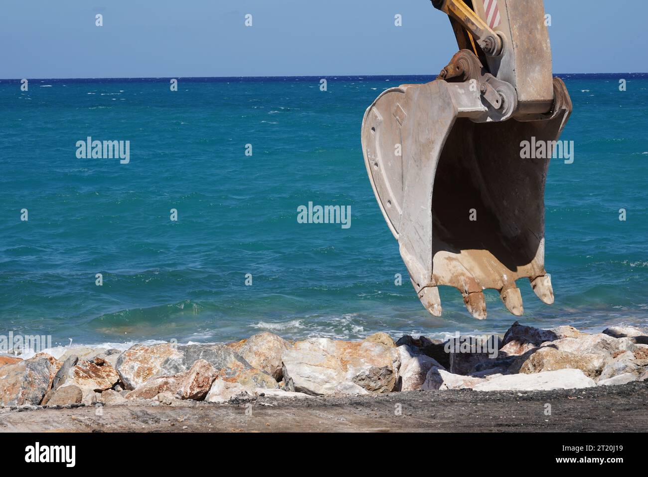 Nahaufnahme einer Löffelschaufel eines Raupenbaggers, der auf der Baustelle arbeitet. Dahinter befindet sich ein Felsenstrand und das Meer von Kreta. Stockfoto