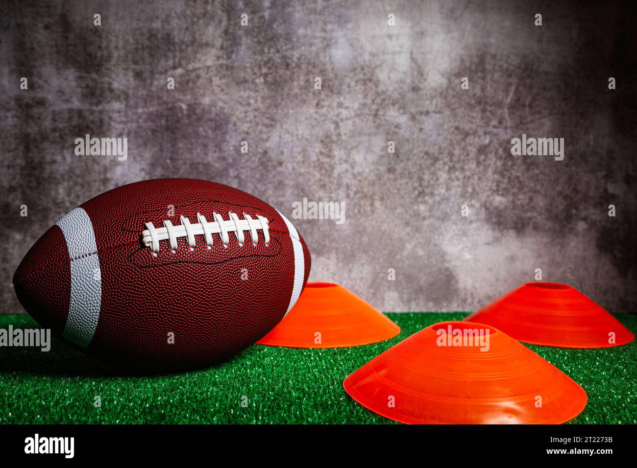 Ein american Football Ball und drei orangefarbene Kegel über grünem Grasboden vor einer grauen Betonmauer. Stockfoto