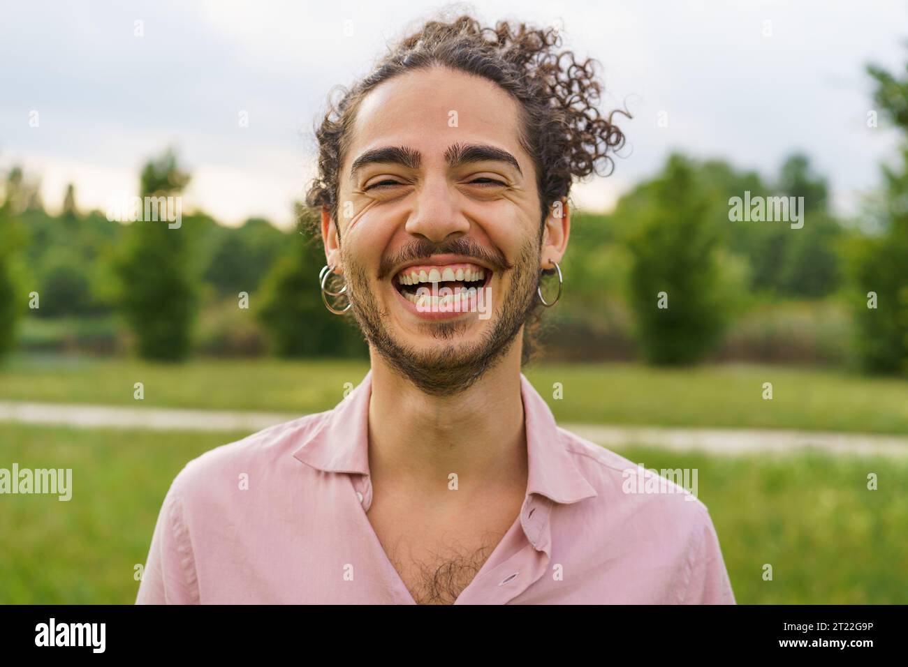 Außenporträt eines jungen Mannes mit lockigen Haaren, die zurückgebunden sind, und lachend mit den Augen. Er trägt Ohrringe und einen Bart. Die Aufnahme, aufgenommen Stockfoto