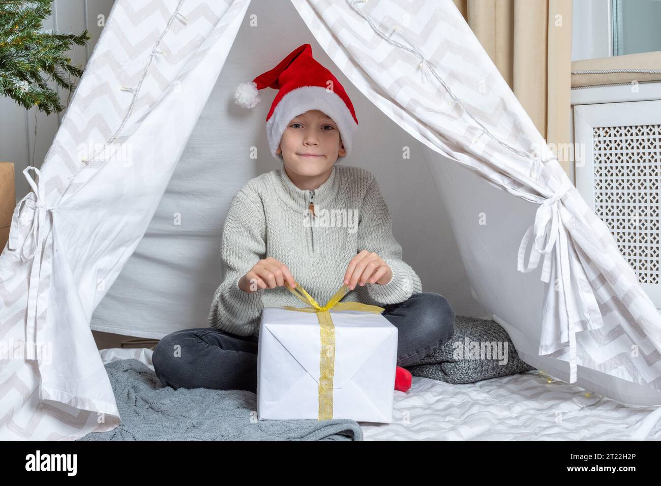 Ein Junge mit Weihnachtsmütze öffnet sein Geschenk zu Weihnachten Neujahr, liegend in einem Kinderzelt Wigwam im Kinderzimmer. Weihnachtsbaum mit Lichtern, Weihnachtszimmer Stockfoto