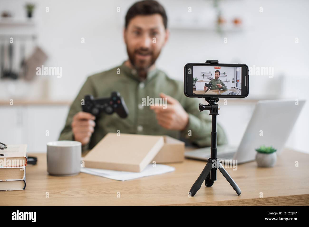 Überglückter kaukasischer Influencer mit modernem Joystick in den Händen, der beim Livestreaming neues Paket entpackt. Online-arbeitender Mann mittleren Alters filmt von Fixed Stockfoto