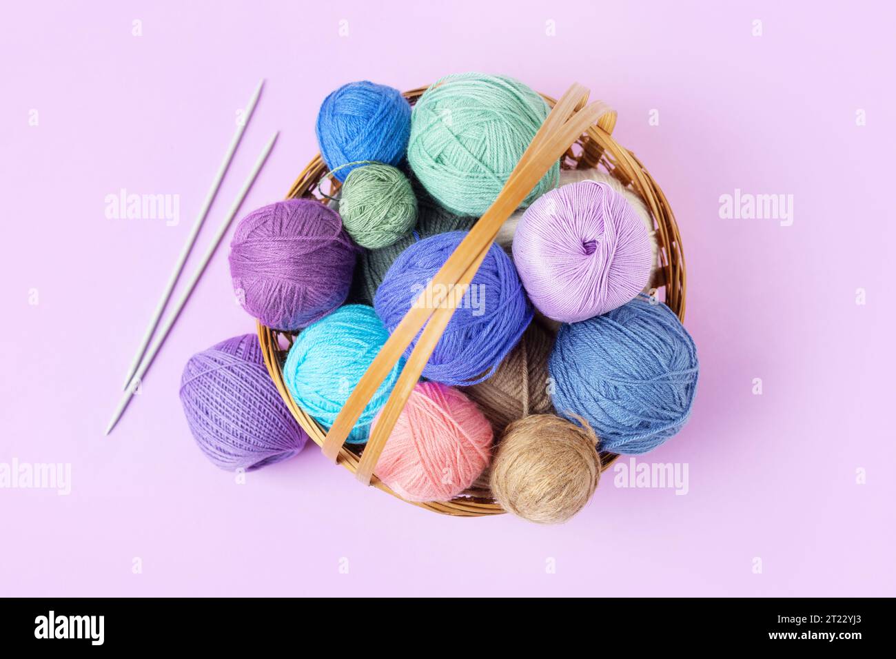 Viele bunte Strickbälle in einem Korb mit Stricknadeln auf einem pastellfarbenen lila Hintergrund. Set für das Hobbys, warme Kleidung oder Spielzeug zu stricken. C Stockfoto