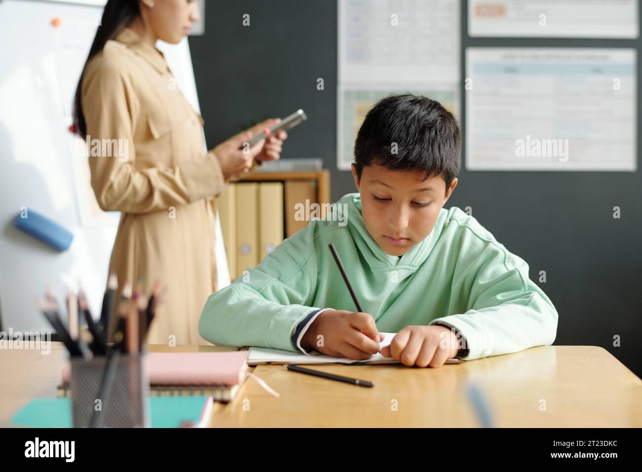 Konzentrieren Sie sich auf clevere und fleißige Schüler, die neue englische Grammatikregeln im Copybook aufschreiben, während Sie am Schreibtisch mit dem Tablet gegen den Lehrer sitzen Stockfoto