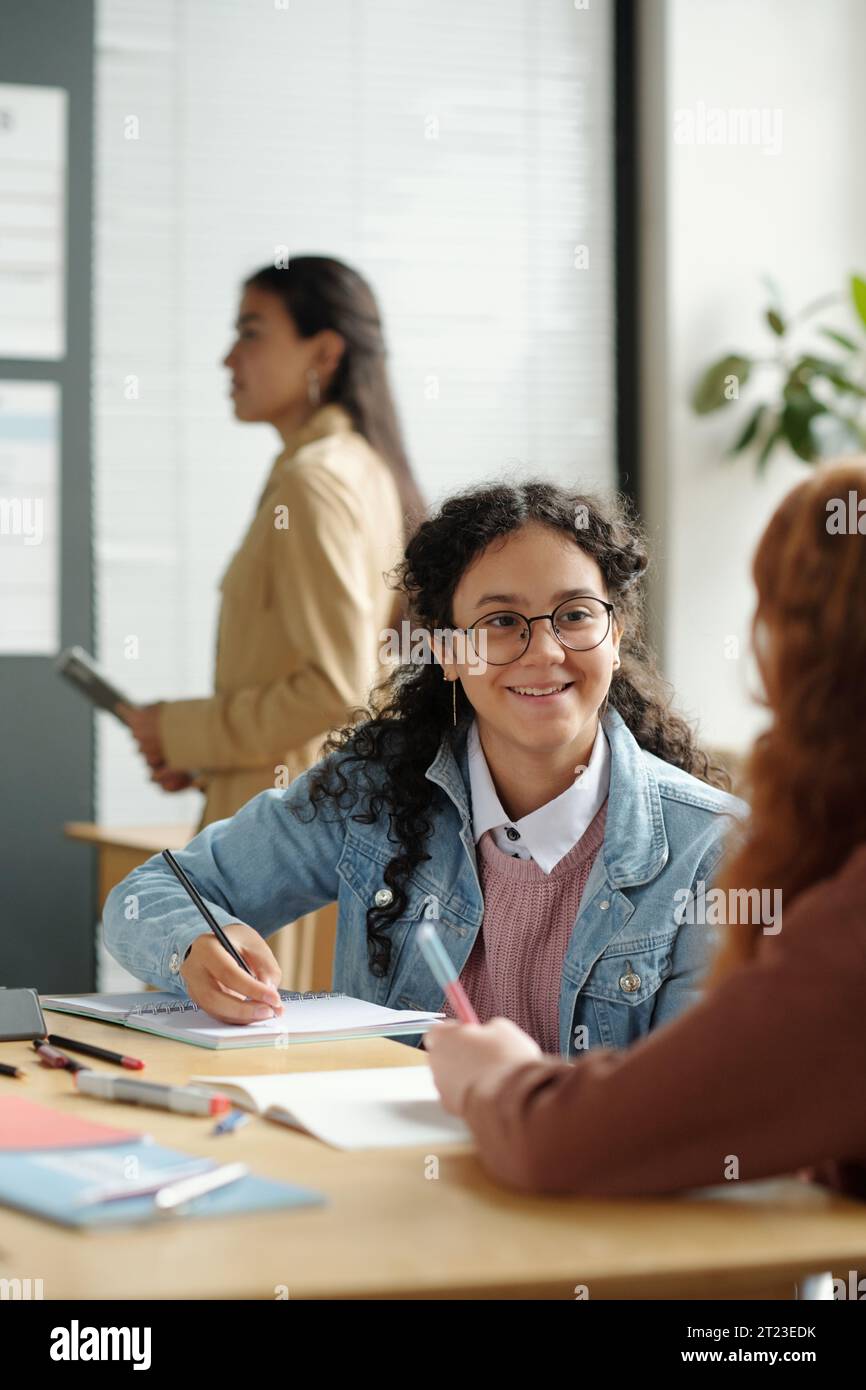 Niedliches lächelndes Schulmädchen mit Stift und Copybook, das den Klassenkameraden beim Englischunterricht während der Diskussion neuer Grammatikregeln ansieht Stockfoto
