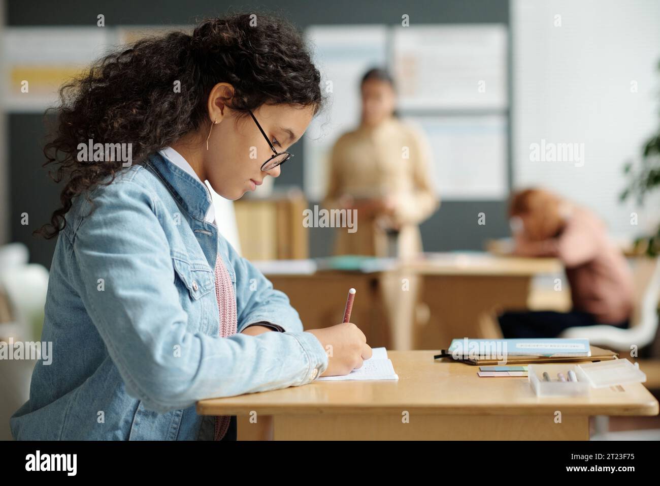 Seitenansicht eines seriösen Schulmädchens in Brille, das Notizen im Copybook macht, während er am Schreibtisch sitzt und an Grammatik der englischen Sprache unterrichtet Stockfoto