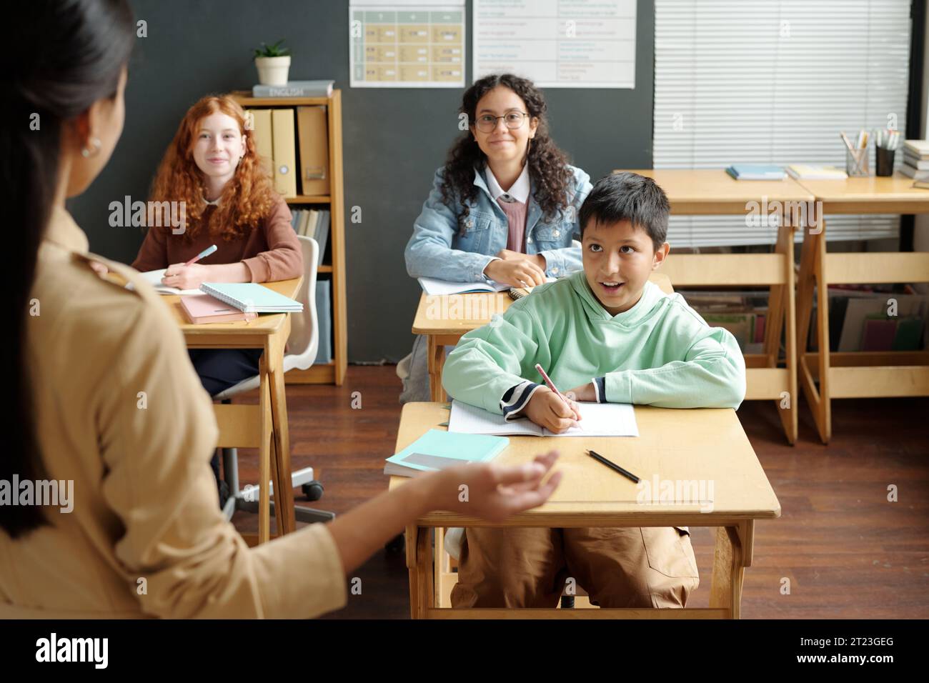 Glücklicher multiethnischer Schuljunge und zwei Schulmädchen, die den Lehrer anschauen und neue Grammatikregeln erklären, während sie vor dem Publikum stehen Stockfoto