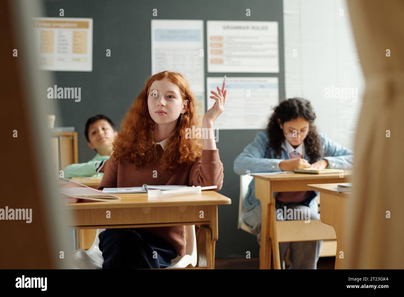 Kluges Schulmädchen hebt die Hand, während es am Schreibtisch sitzt und auf das Whiteboard blickt, während es über neue Regeln der englischen Grammatik spricht Stockfoto