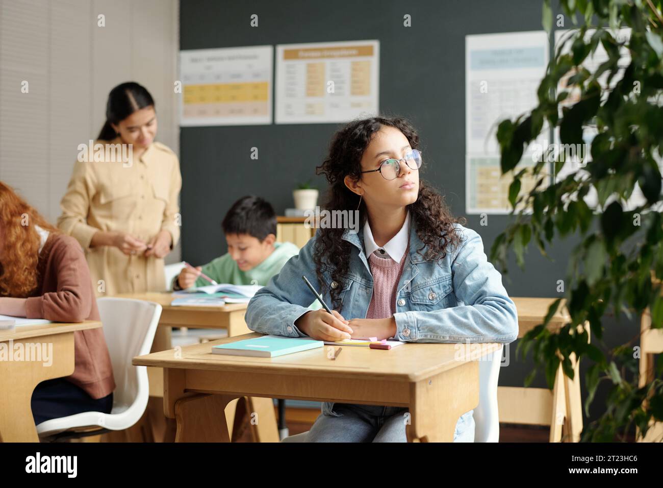 Niedlicher nachdenklicher Schüler der Sekundarstufe, der durch das Fenster des Klassenzimmers schaut, während er am Schreibtisch sitzt und einen Essay im Unterricht aufschreibt Stockfoto