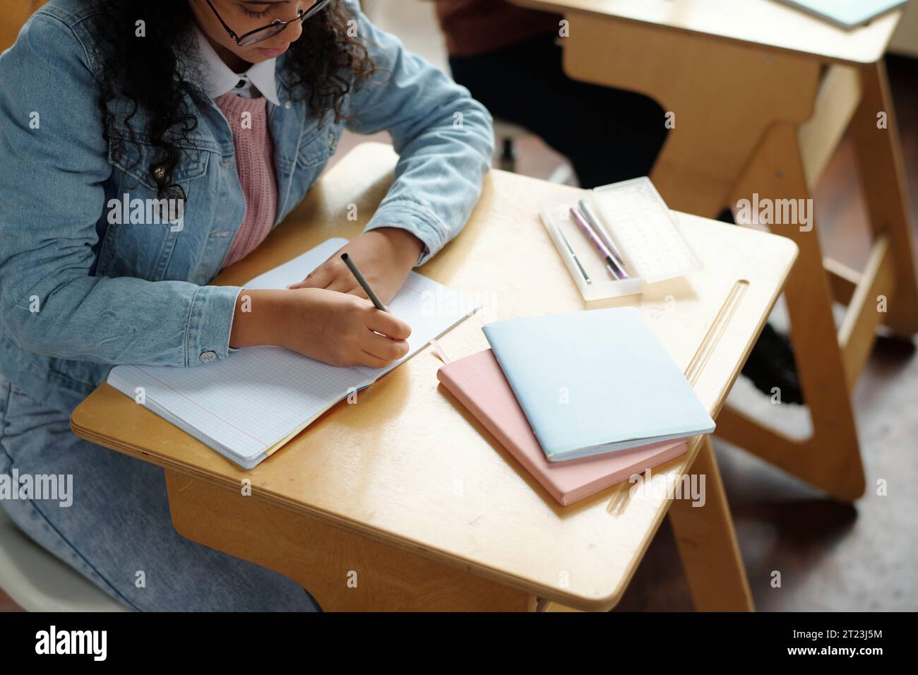 Über dem Winkel eines fleißigen multiethnischen Schulmädchens, das beim Unterricht Notizen auf einer leeren Seite des Copybook macht, während er mit Notizbüchern und Stiften am Schreibtisch sitzt Stockfoto
