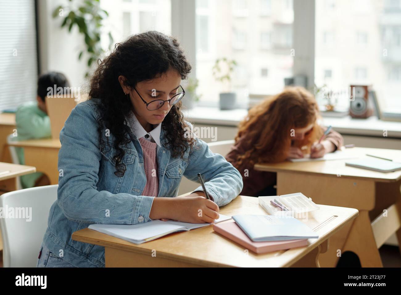 Hübscher multiethnischer Schüler in Casualwear, der Notizen im Copybook macht, während er vor der Kamera gegen Klassenkameraden sitzt Stockfoto