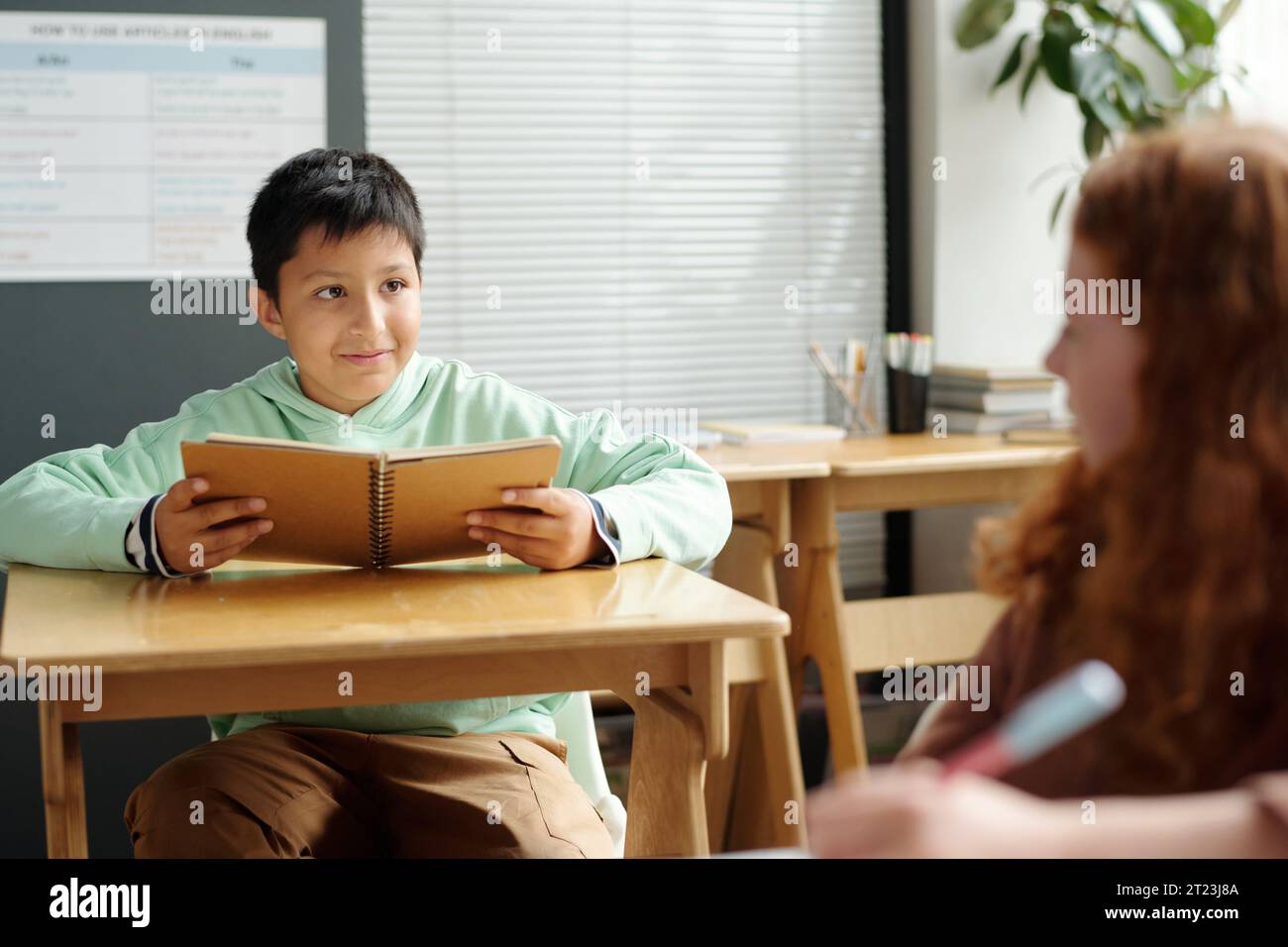 Niedlicher Schuljunge mit offenem Copybook, der seinen Klassenkameraden beim Unterricht ansieht, während er am Schreibtisch sitzt und sich über einige Grammatikpunkte berät Stockfoto