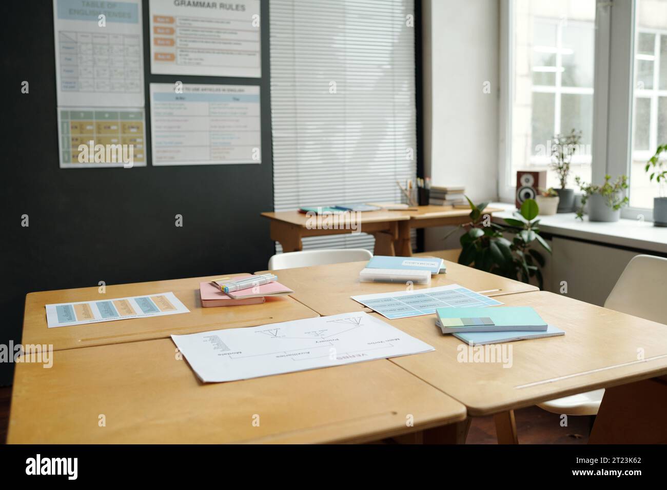 Teil des geräumigen Klassenzimmers mit quadratischem Holzschreibtisch, der in der Mitte vor dem Fenster steht, Wand mit Grammatiktischen, grünen Pflanzen und anderen Dingen Stockfoto