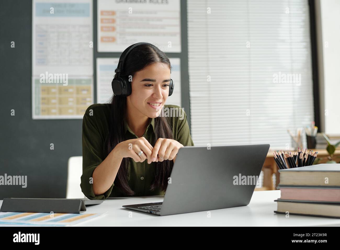Junger, lächelnder Lehrer oder Schüler eines Englischkurses in Kopfhörern, der am Schreibtisch vor dem Laptop sitzt und auf den Bildschirm blickt Stockfoto