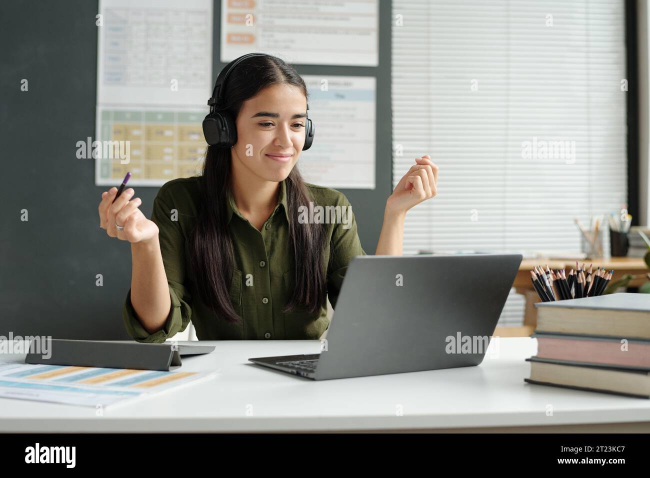 Selbstbewusste junge Lehrerin in Kopfhörern, die Online-Lernenden Grammatikregeln erklärt, während sie am Arbeitsplatz vor dem Laptop-Bildschirm sitzt Stockfoto