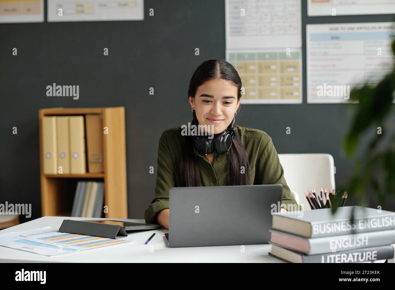 Glückliche junge Schülerin, die online Englischunterricht ansieht, während sie am Schreibtisch vor dem Laptop sitzt und auf den Bildschirm blickt Stockfoto