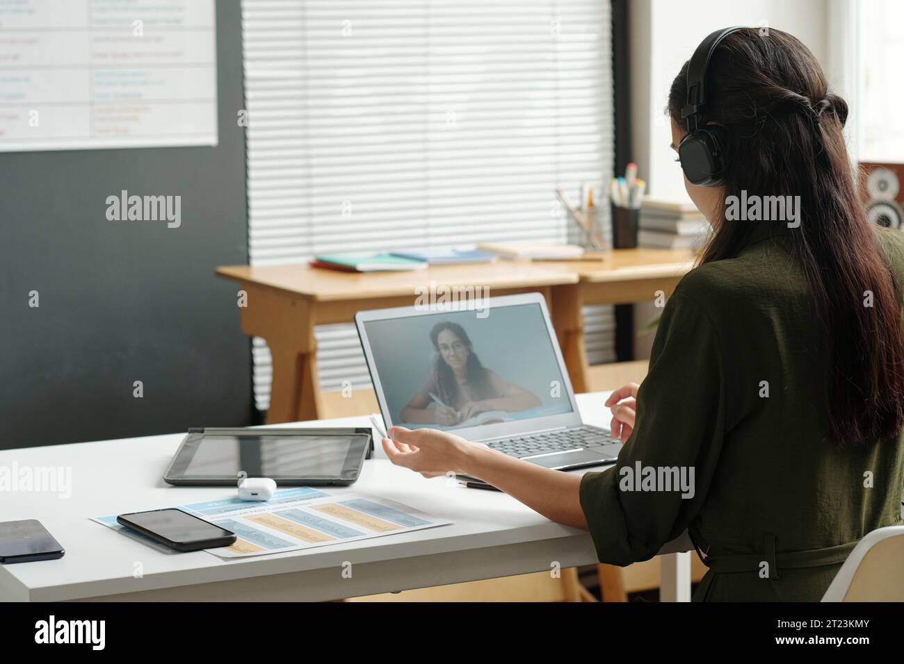 Rückansicht einer jungen Lehrerin in Kopfhörern, die das Schulmädchen auf dem Laptop-Bildschirm ansieht und ihr einige neue Regeln der englischen Grammatik erklärt Stockfoto