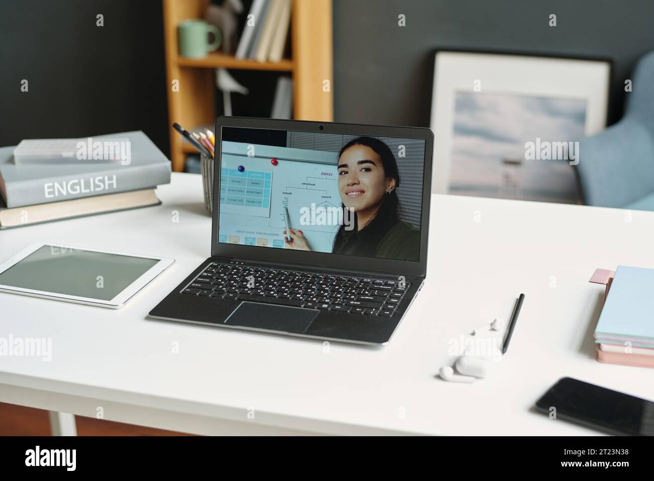 Laptop mit Englischlehrer auf dem Bildschirm, auf dem Schreibtisch mit Tablet, Stapel Bücher, Wörterbücher, Kopfhörer, Bleistift und Handy des Schulkindes Stockfoto