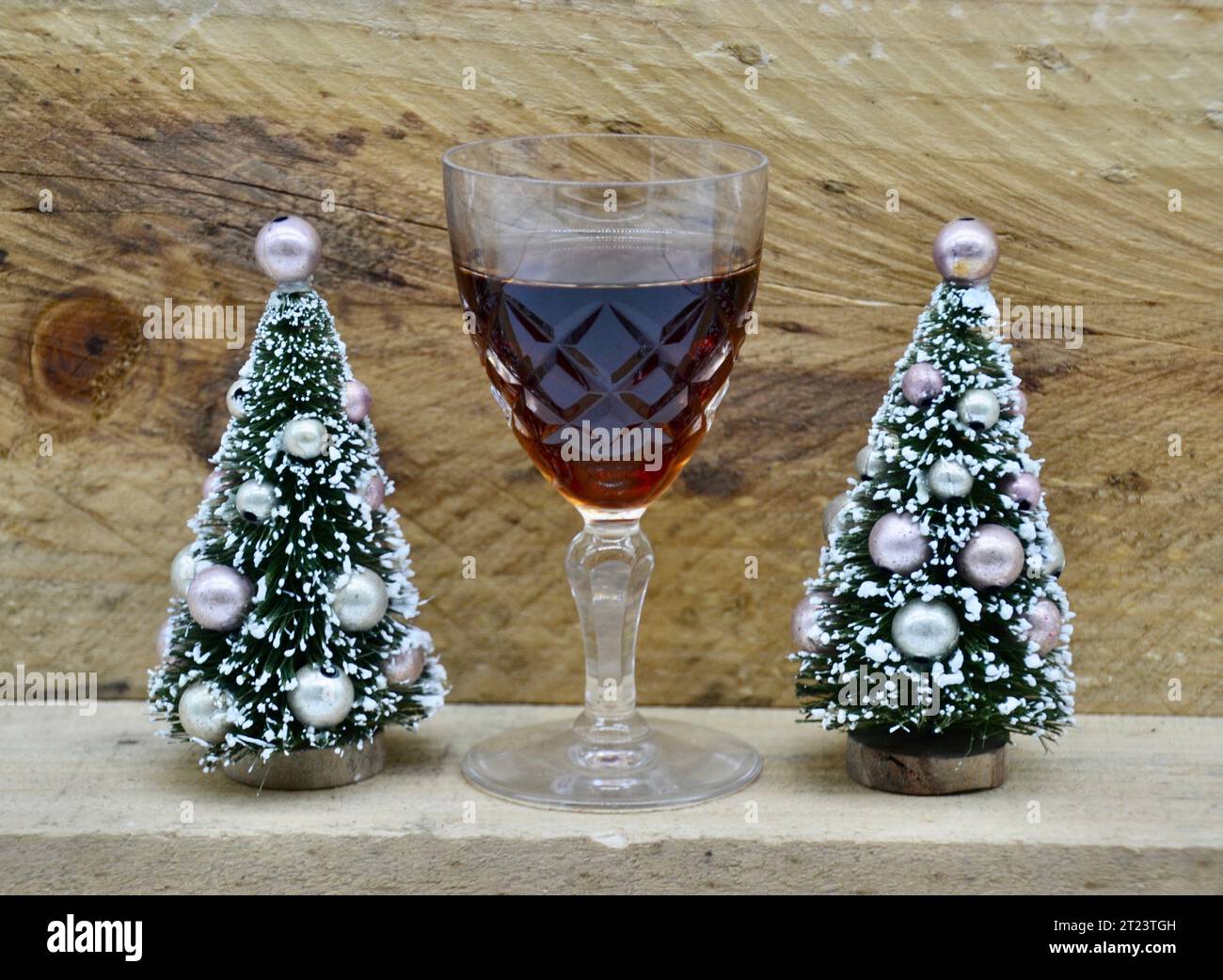 Zwei kleine verzierte, gefälschte, schneebedeckte Weihnachtsbäume mit einem Glas Portwein oder Sherry für den Weihnachtsmann vor einem rauen Holzhüttenhintergrund Stockfoto