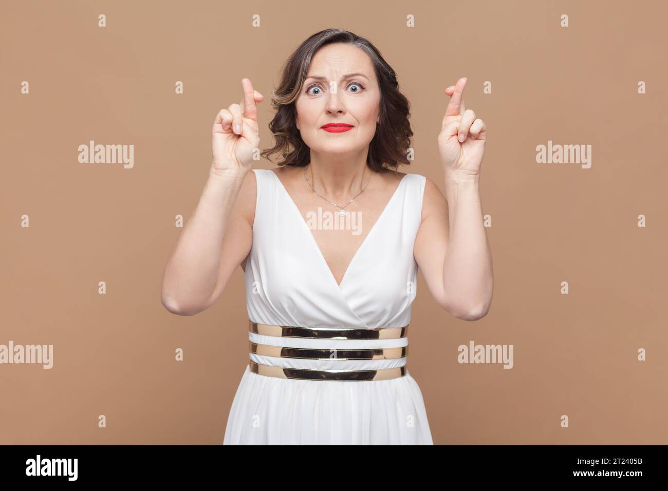 Porträt einer angenehm aussehenden hoffnungsvollen Frau mittleren Alters mit gewelltem Haar kreuzt die Finger und hat große Hoffnung auf ein besseres, weißes Kleid. Innenstudio, isoliert auf hellbraunem Hintergrund. Stockfoto