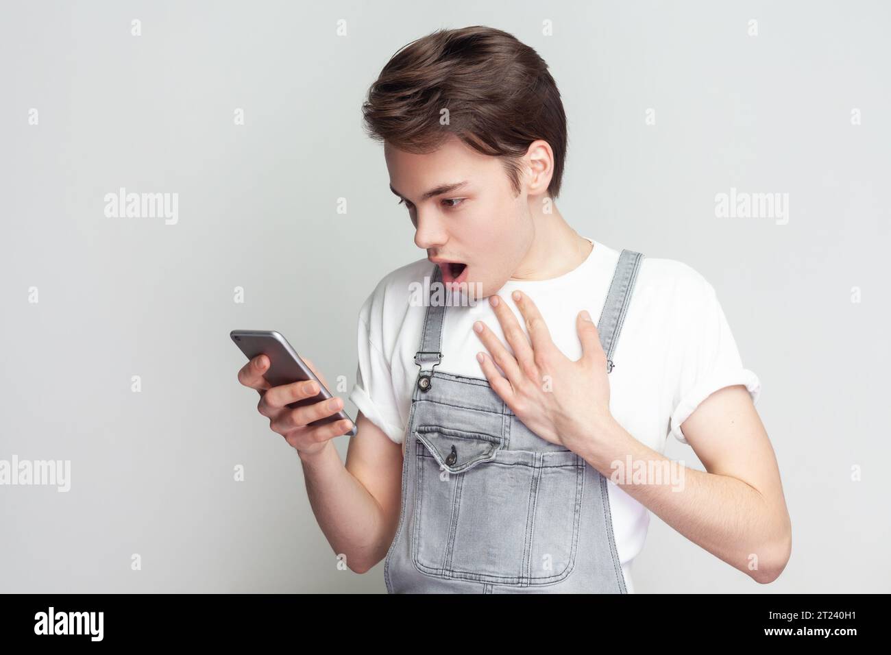 Porträt eines schockierten, staunenden jungen Brünetten, der mit dem Handy steht, schockierende Nachrichten liest, überrascht ist und Denim-Overalls trägt. Innenstudio isoliert auf grauem Hintergrund. Stockfoto
